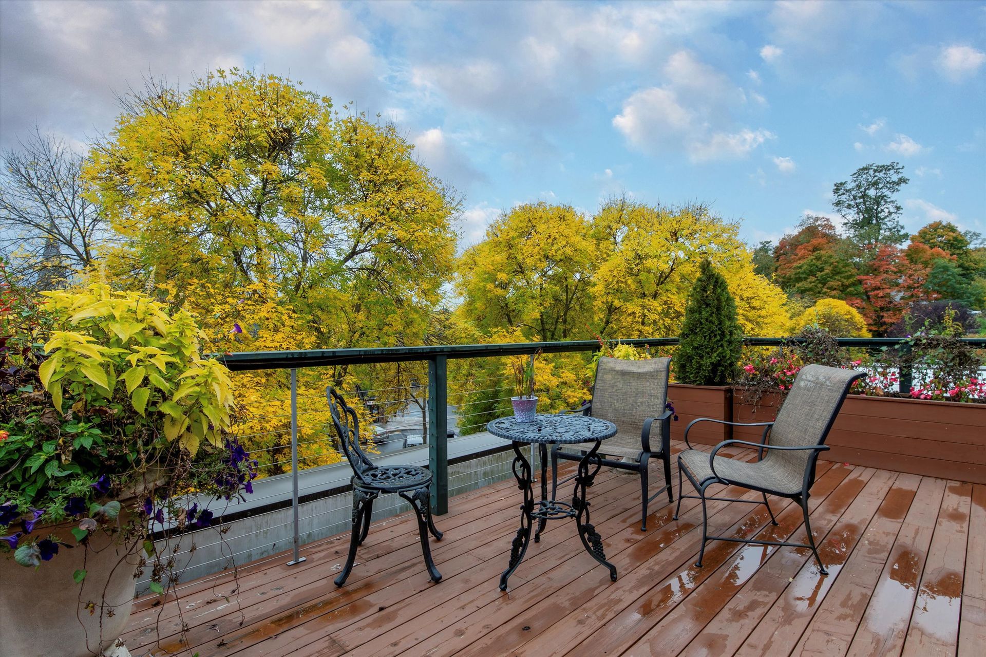 A small bistro table and chairs on a wooden deck overlooking autumn trees under a cloudy sky.