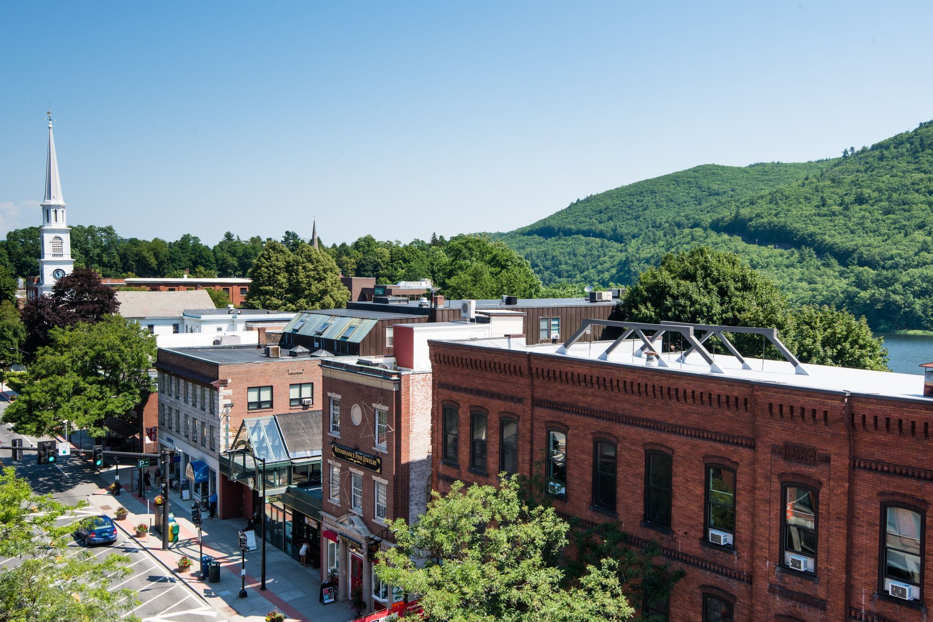 A scenic view of a sunny downtown street lined with historic red brick buildings, a church steeple, and green hills.