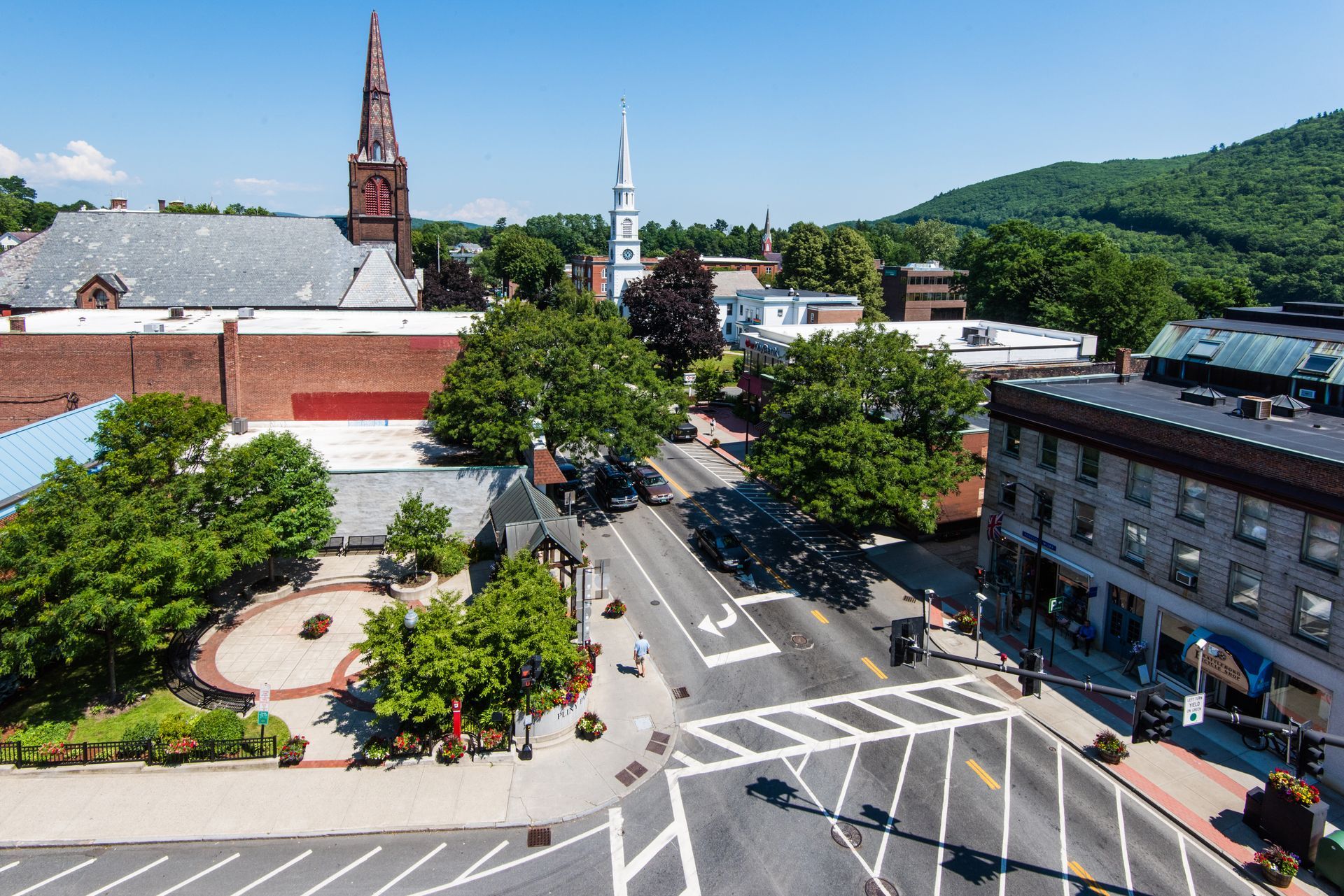 An aerial view of a town square featuring a brick building, two white church steeples, trees, and a tree-lined street.