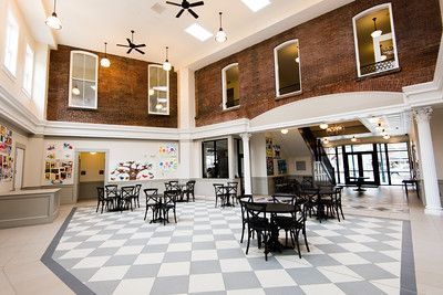 An open, airy atrium with a checkerboard floor, round tables with black chairs, brick walls, and high ceiling windows.