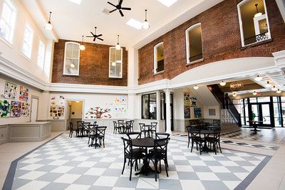 A bright, spacious indoor hall with high ceilings, brick walls, a checkered floor, and multiple tables and chairs.