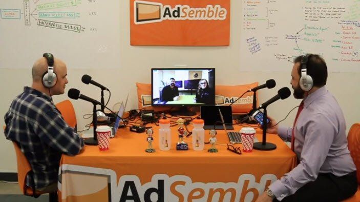 Two men are sitting at a table with microphones in front of a sign that says adsemble