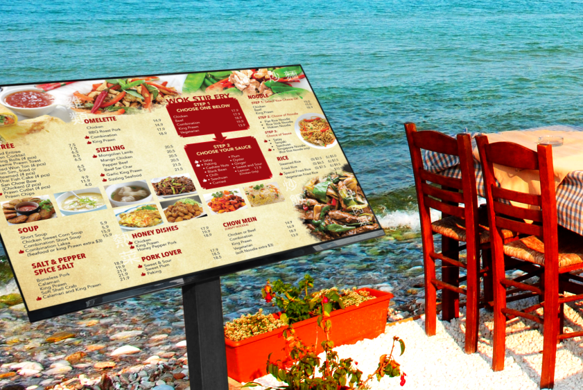 A menu is displayed on a sign next to a table and chairs by the ocean.