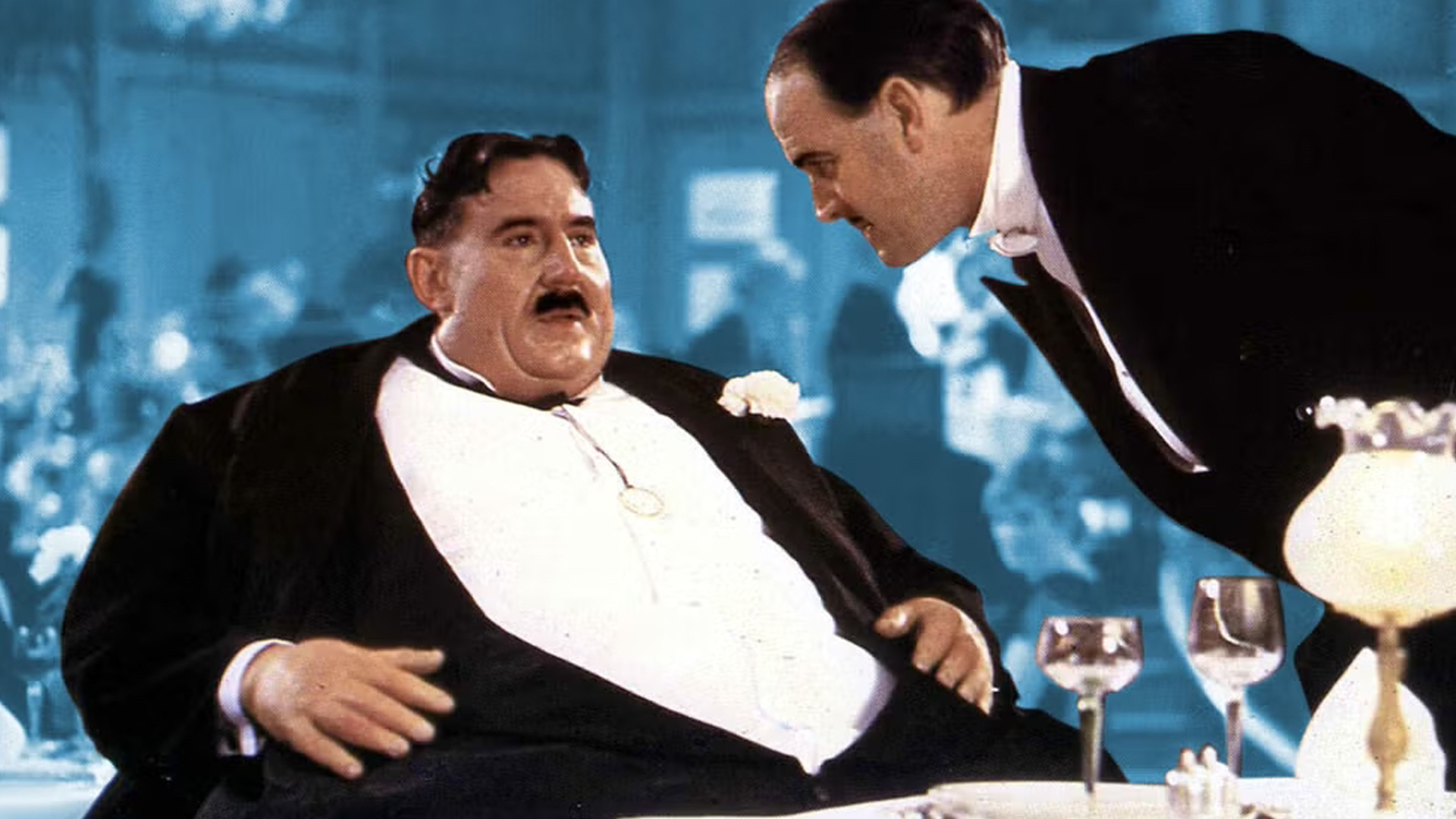 A man in a tuxedo is sitting at a table talking to an enormous customer.