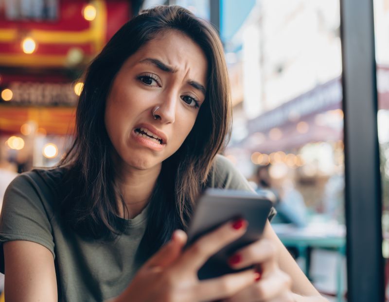Woman looking worried at a smartphone in a café, with blurred city lights in the background