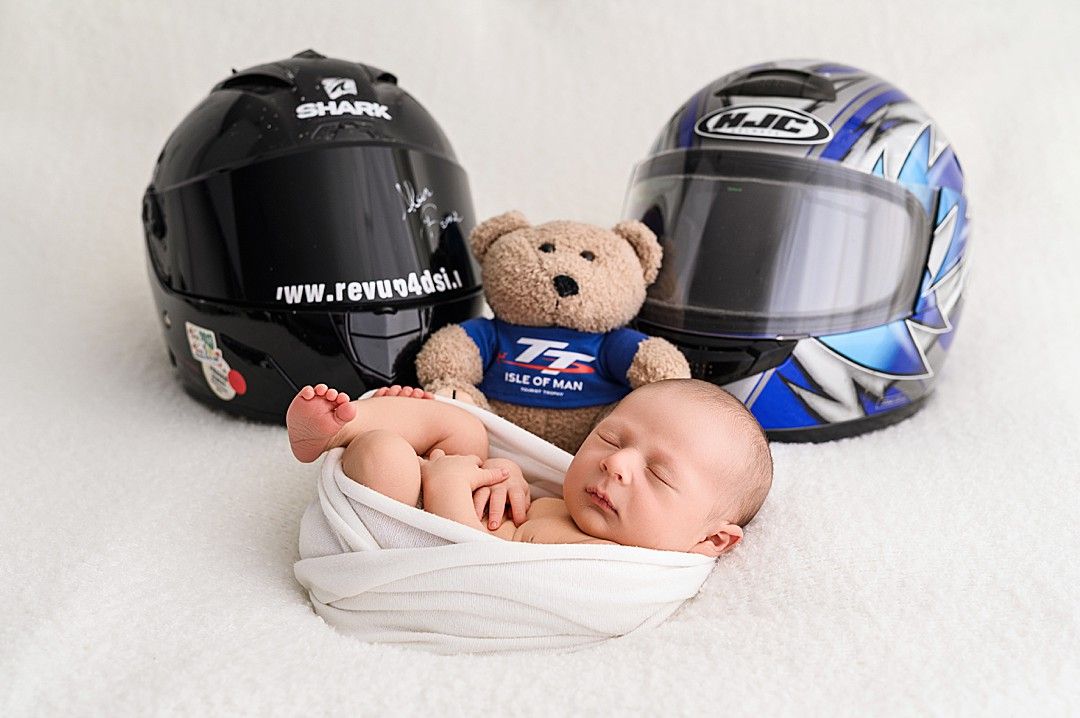 Sleeping newborn wrapped in white swaddle with teddy bear and motorbike helmets in Dublin studio