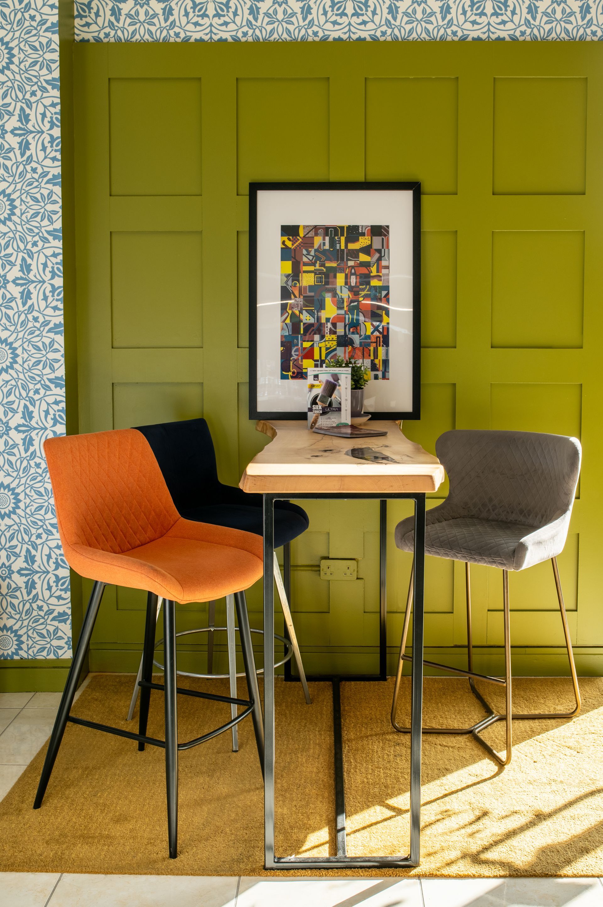 A picture of a green wall with a colourful picture infront on a table with three chairs