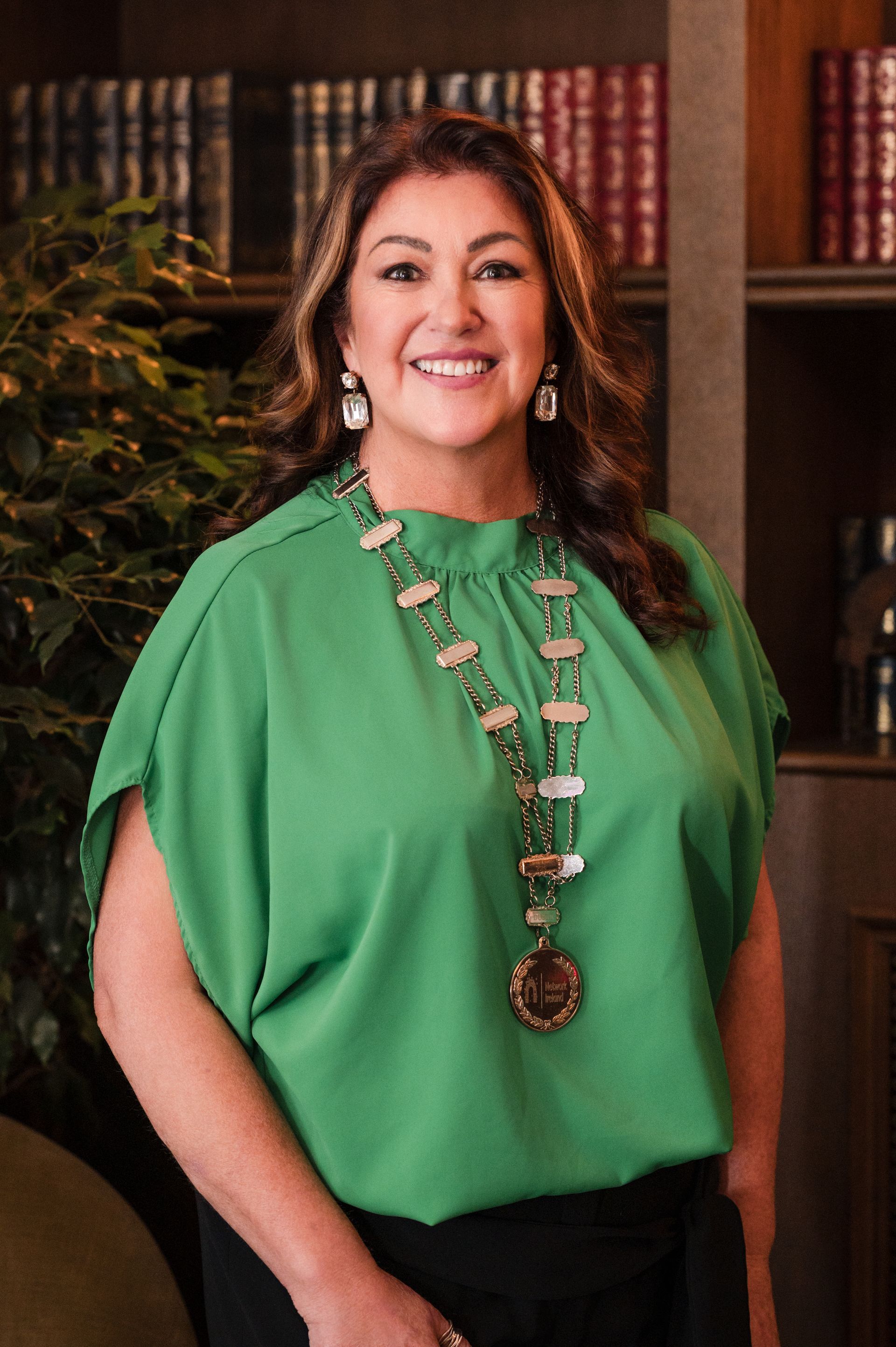 Professional headshot of a businesswoman smiling indoors in a Dublin venue, wearing a green blouse and statement jewellery, with bookshelves in the background.