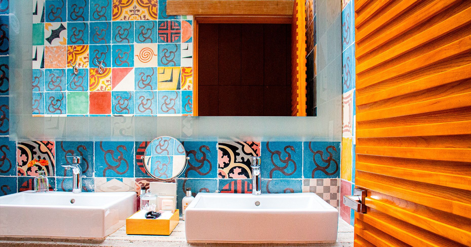 Bathroom with colorful patterned tiles, two white sinks, and a large mirror. Bright orange door on the right.