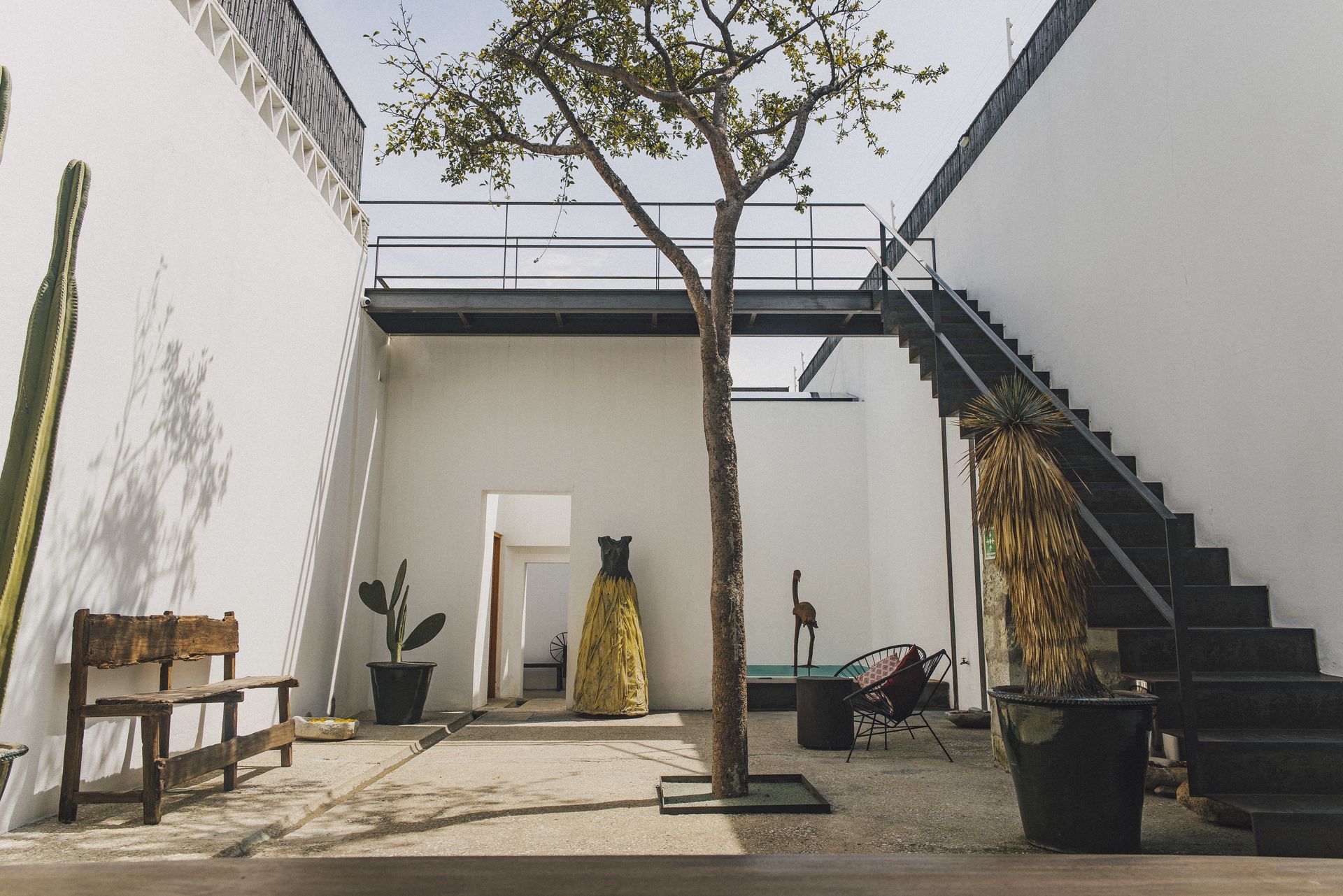 White-walled courtyard with tree, staircase, and bench. A metal bridge extends overhead.