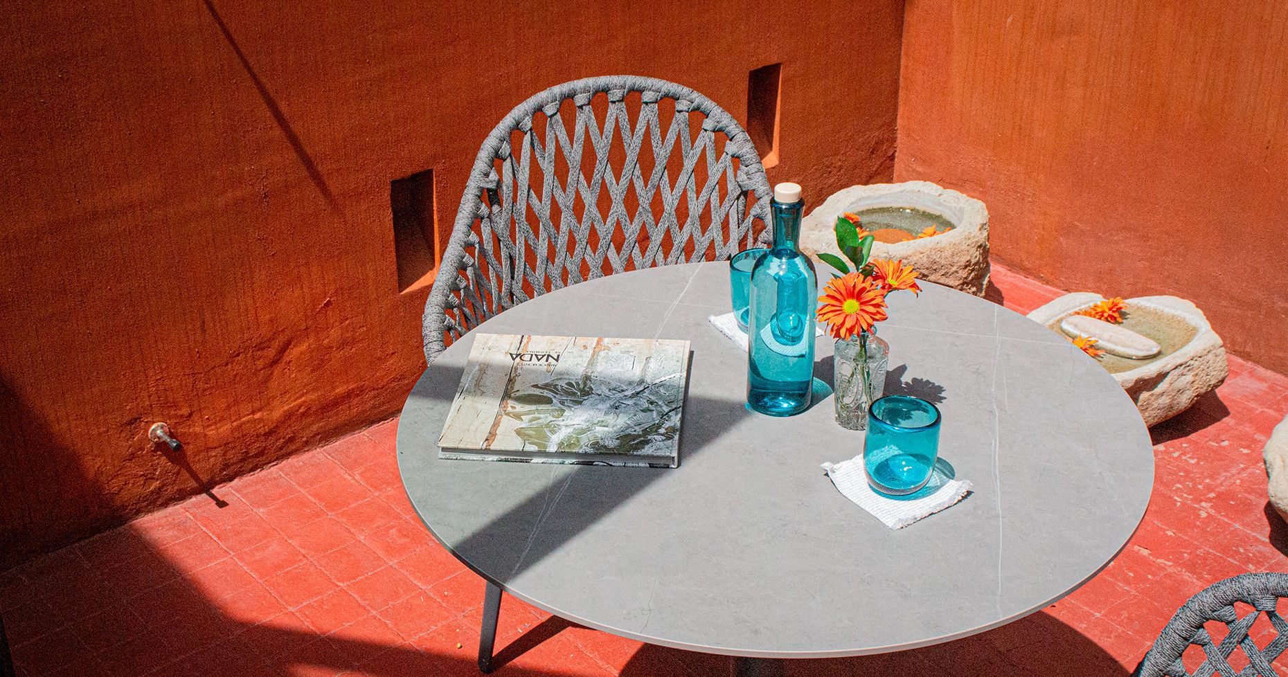 Outdoor patio with round gray table, blue bottle, flowers, and chair against orange wall.