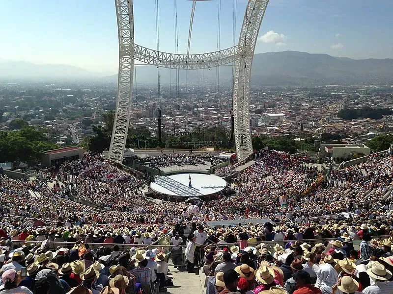 Large crowd in an outdoor amphitheater, stage at the center with a tall structure overhead, cityscape backdrop.