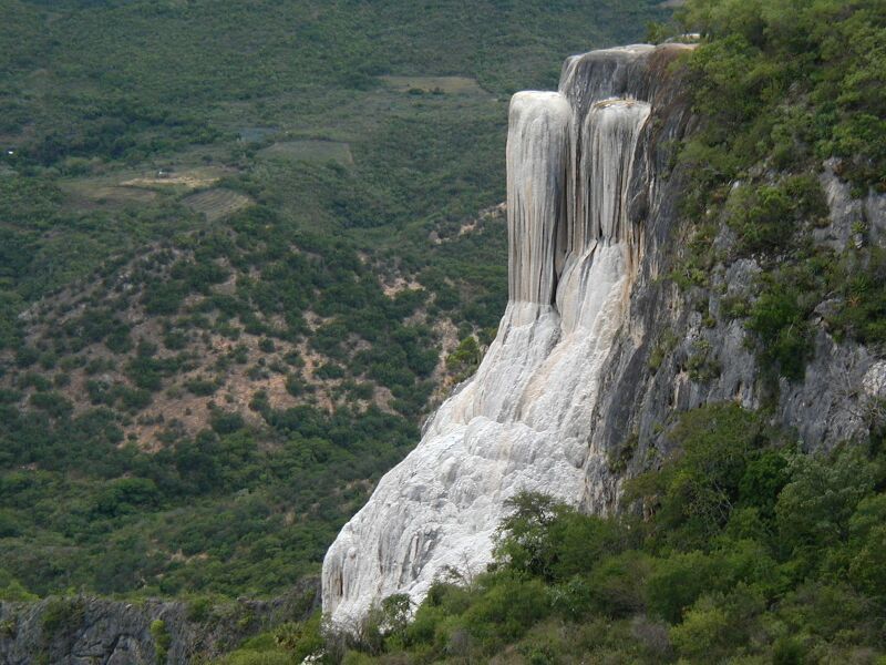 White mineral formations cascading down a cliff face, surrounded by green vegetation and a distant landscape.
