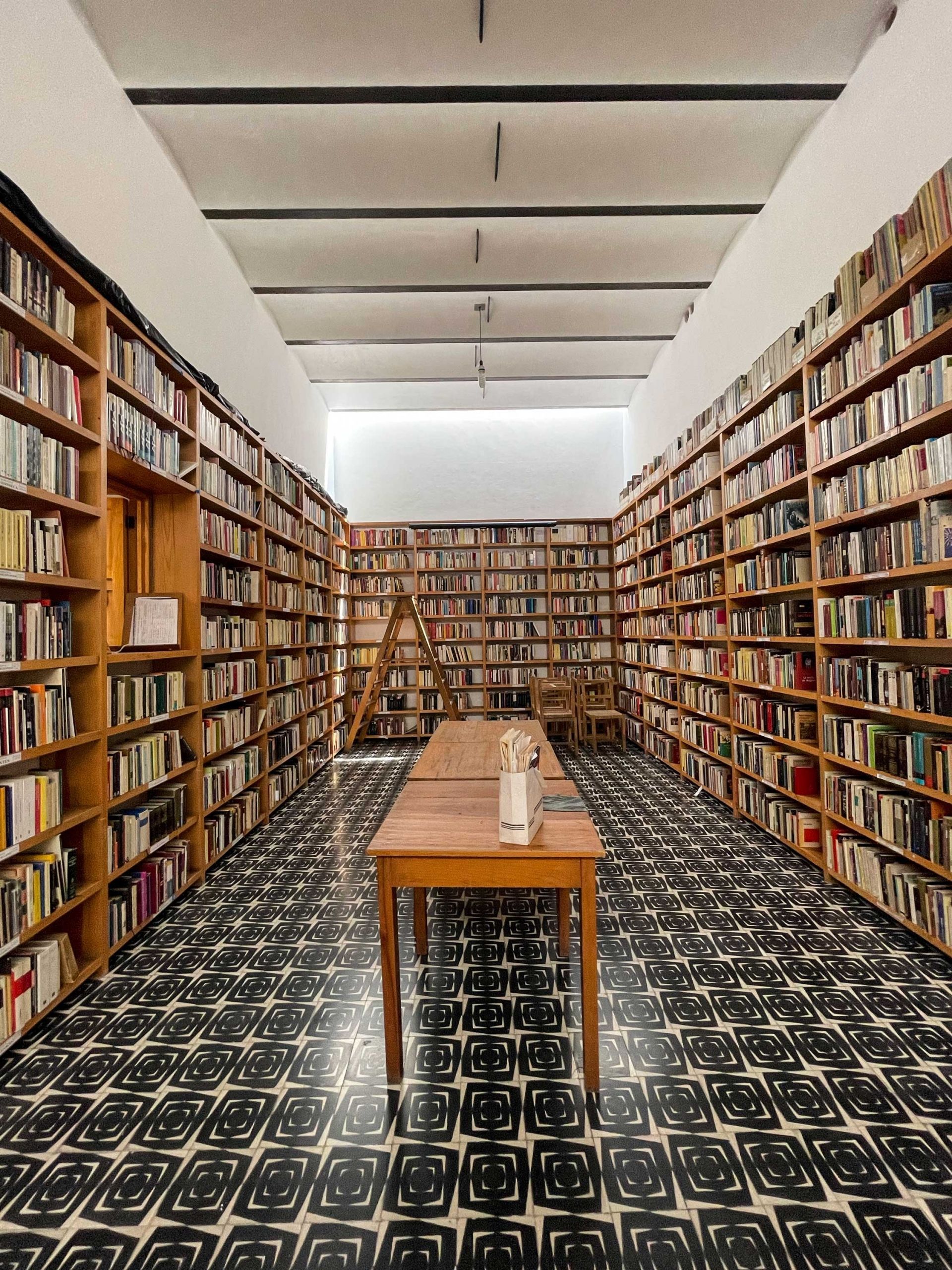 Long, narrow bookstore with floor-to-ceiling bookshelves. A wooden table with flowers sits in the middle.