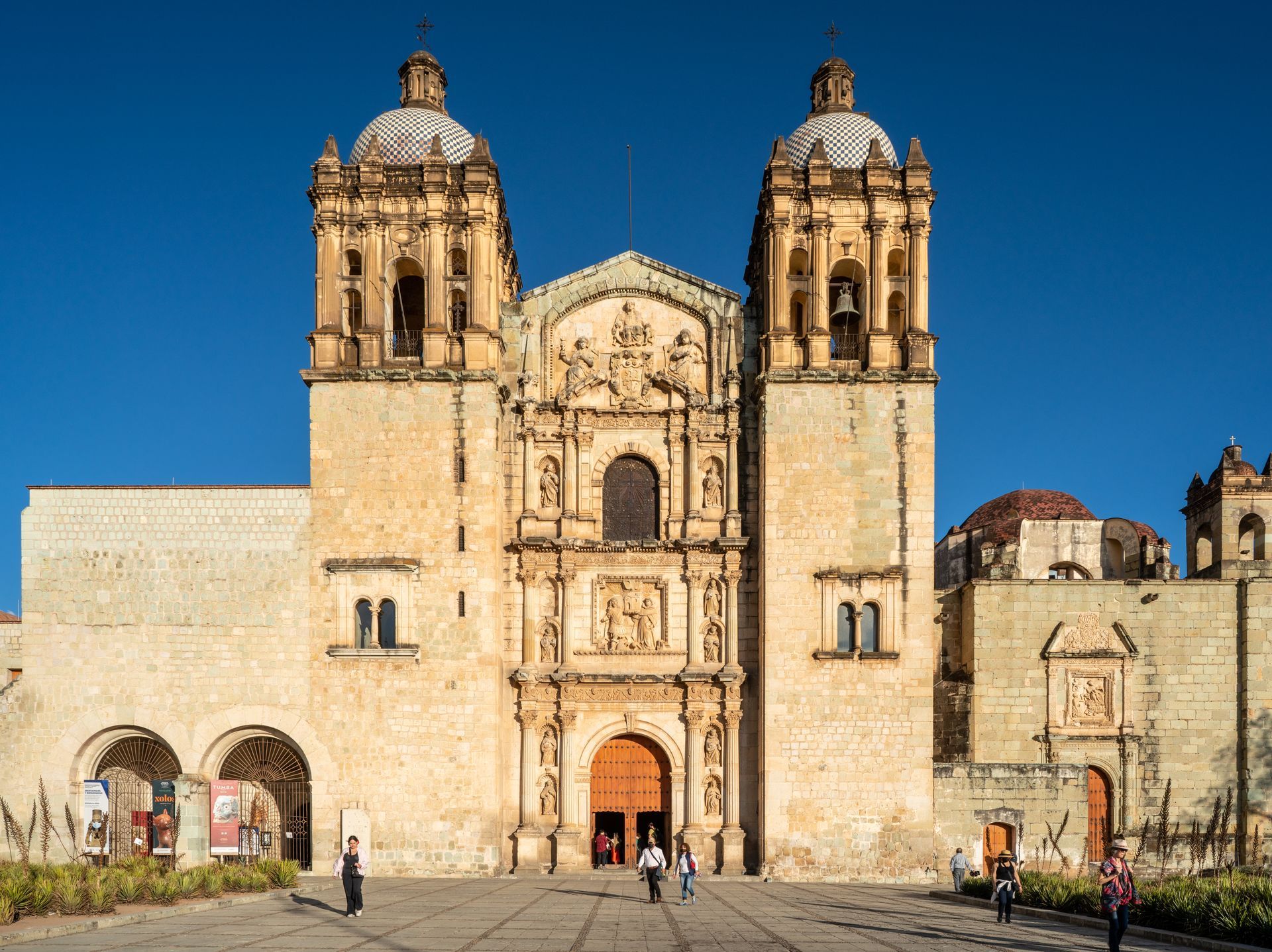 Large, ornate church with two bell towers, beige walls, and a blue sky. People near the entrance.