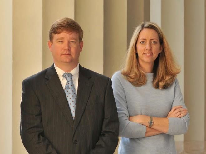 Two people standing before columns, one in a dark suit and one in a light blue top with arms crossed.