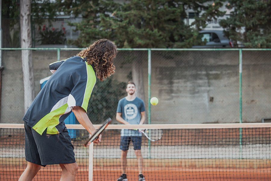 Two men are playing tennis and one has a smiley face on his shirt