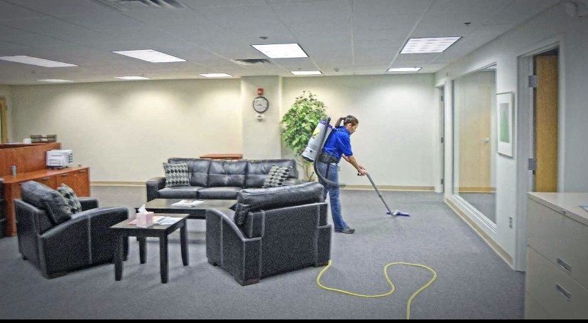 A person wearing a backpack vacuum cleans the carpeted floor of an office lobby furnished with dark leather sofas.
