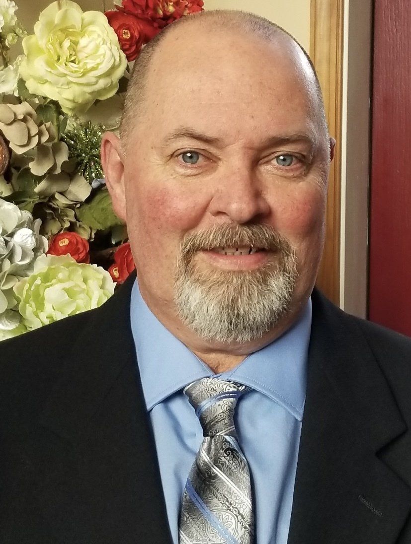 A person wearing a suit, light blue shirt, and patterned tie, posed against a backdrop of soft flowers.