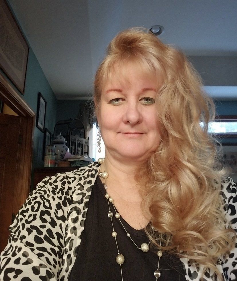 A person with long, blonde, wavy hair wearing a black-and-white leopard print cardigan and a beaded necklace.