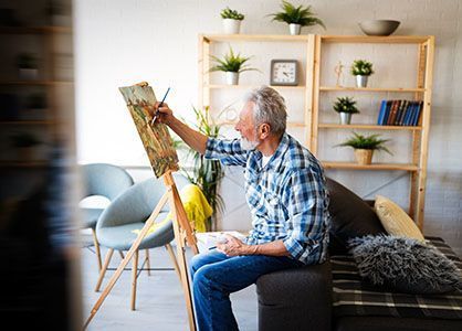 An elderly man is sitting on a couch painting on an easel.