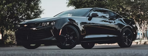 Black Chevrolet Camaro parked on asphalt. Black wheels, dark setting, trees in the background.
