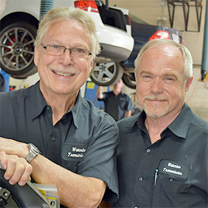 Two men are posing for a picture in front of a car in a garage.