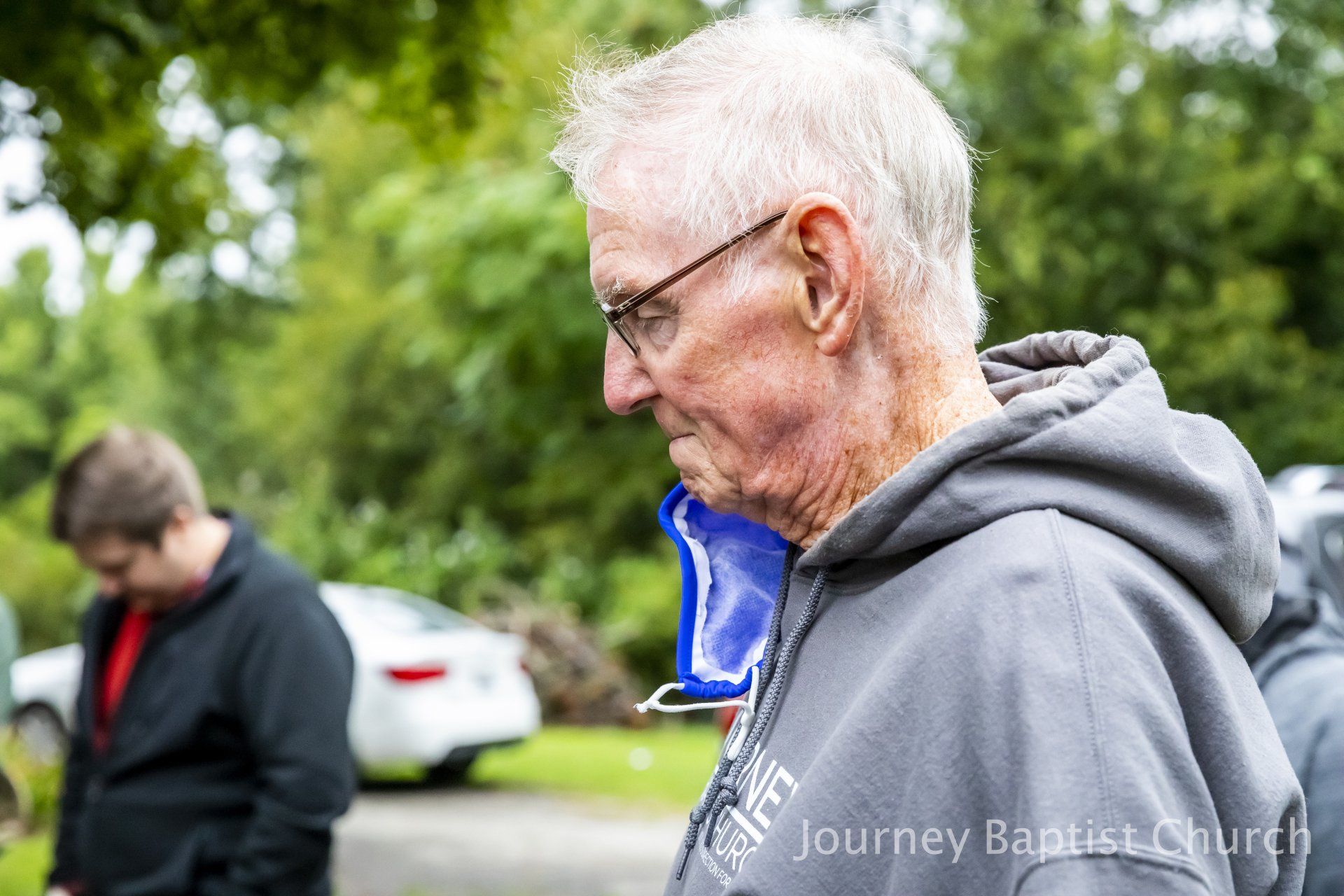 Older man in gray hoodie with glasses, head bowed, outdoors, with a blurred figure in the background.