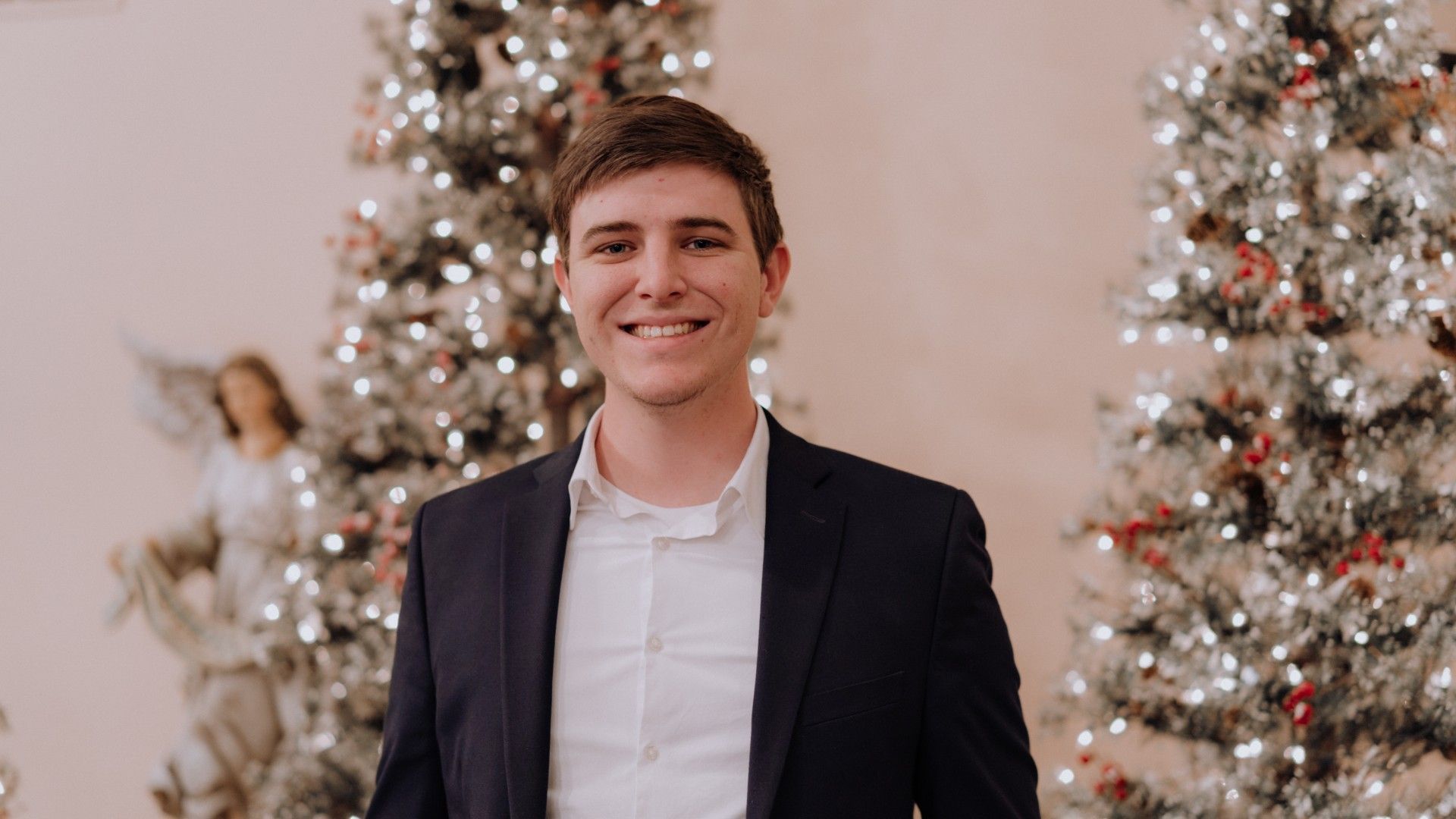 Man in a navy suit smiles in front of Christmas trees and angel statue.