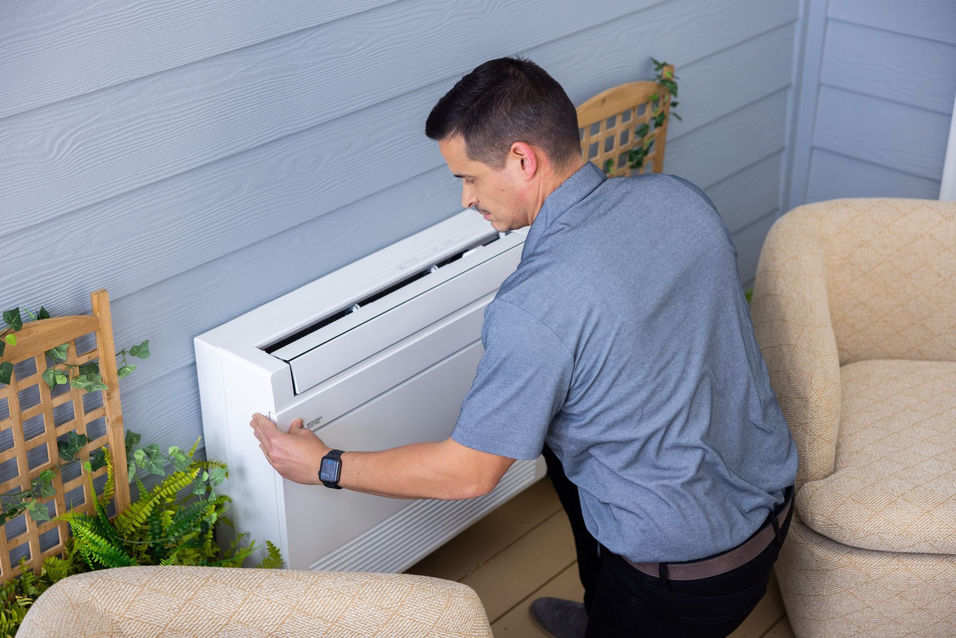 Pfluke Home Services technician installing a white air conditioning unit on a light blue wall, outdoors.
