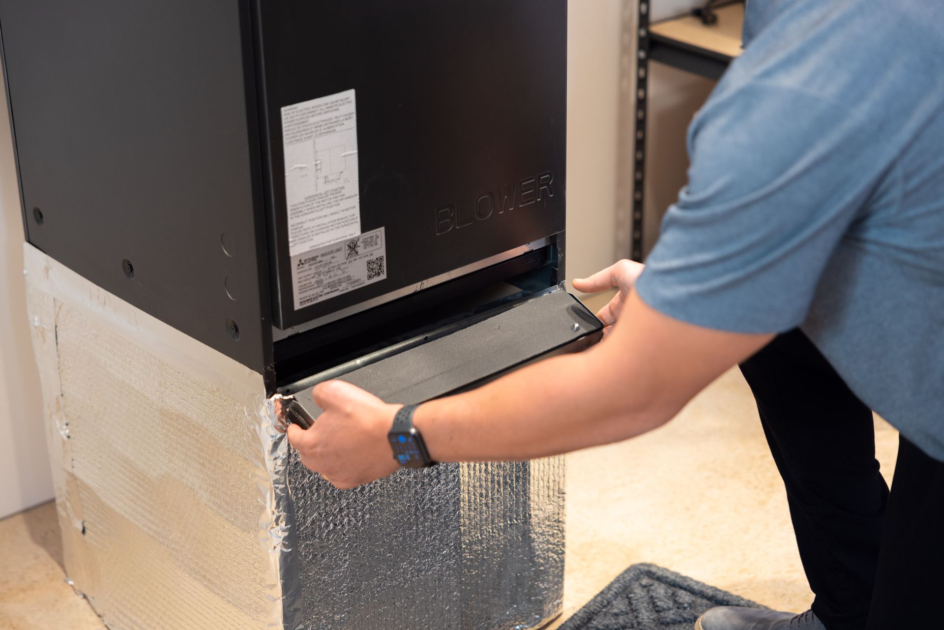 Pfluke Home Services technician installing a black air filter into a furnace. Silver insulation is visible.