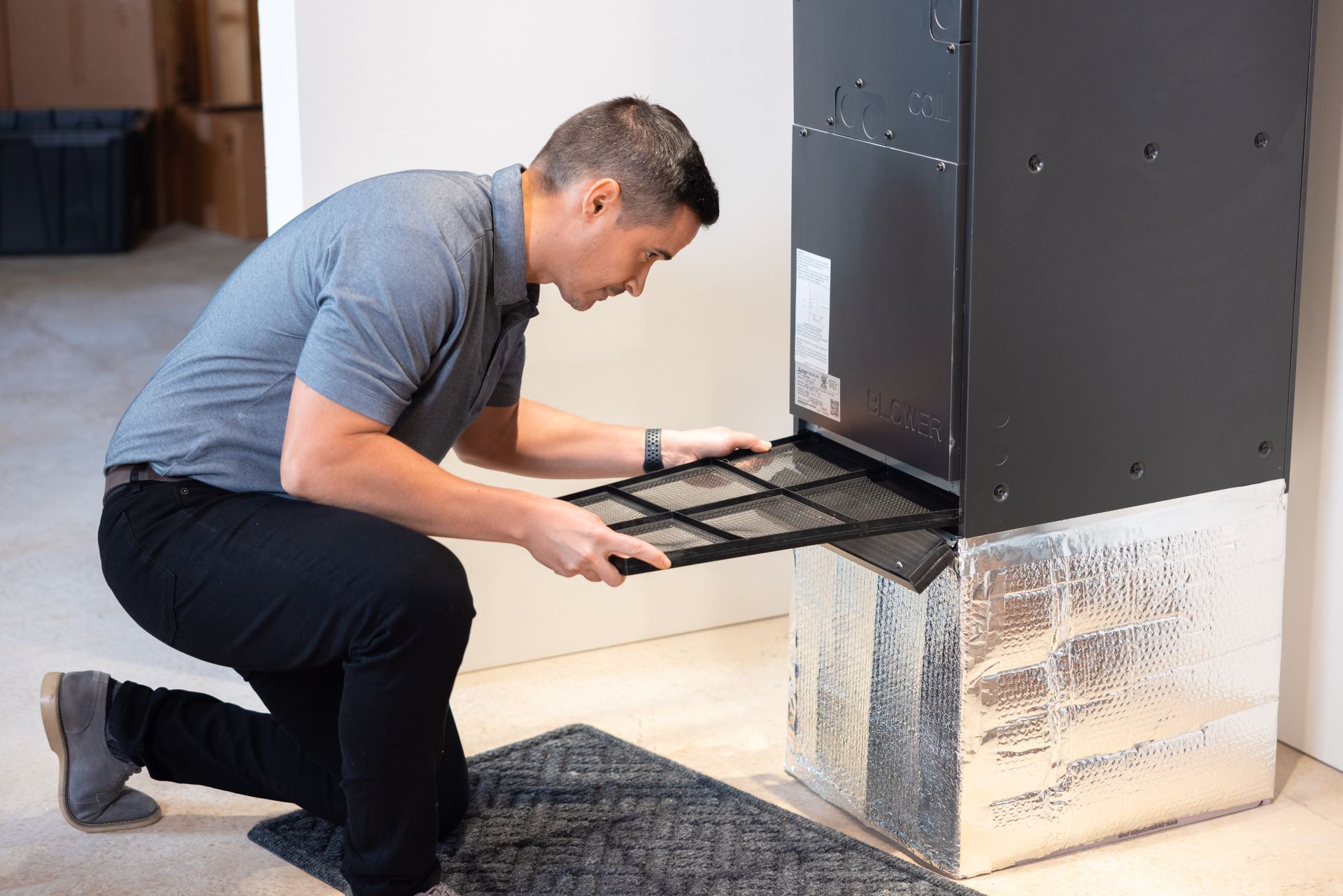 Pfluke Home Services technician kneeling, removing air filter from a black furnace, inside a home. Filter is black.