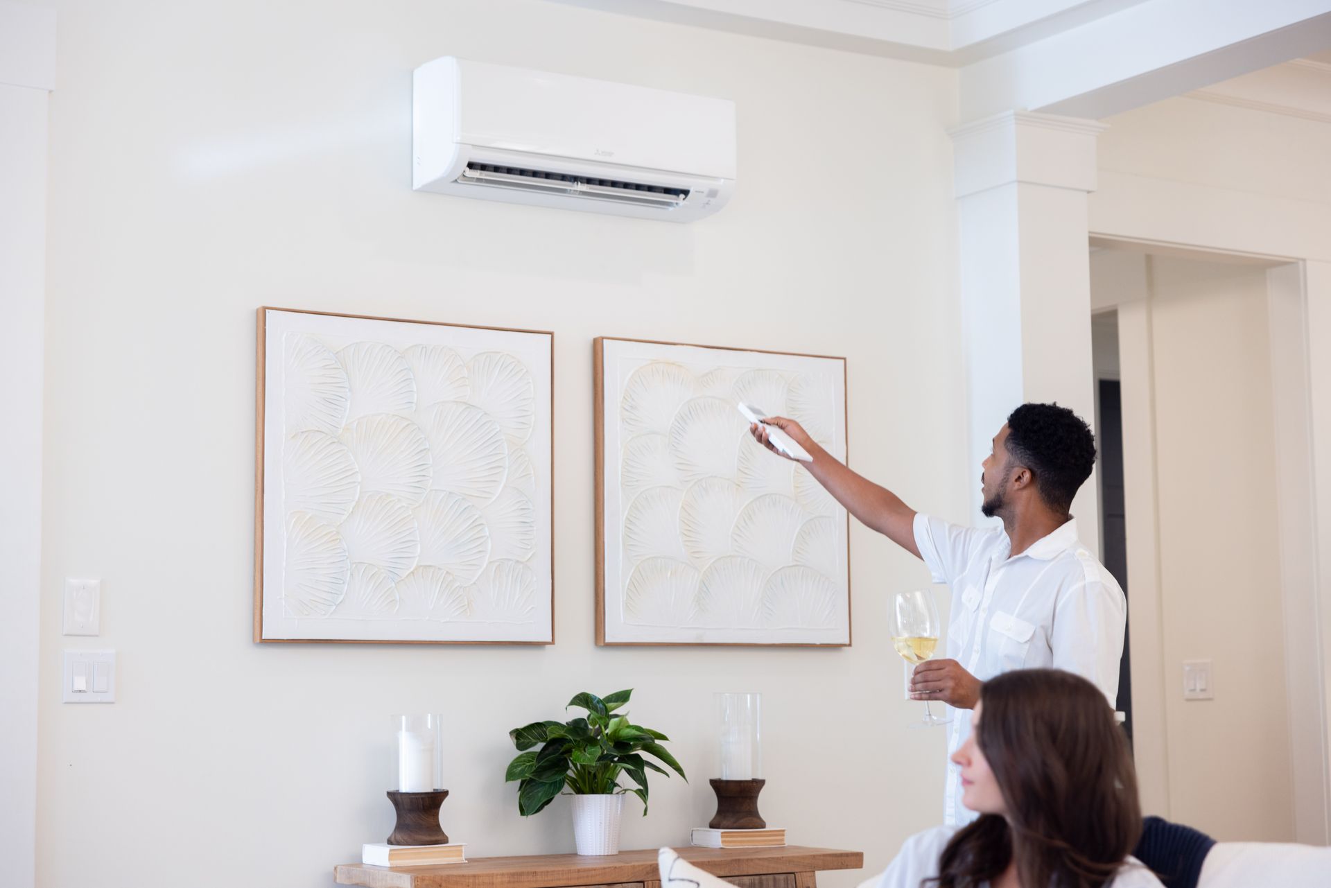 Man adjusting air conditioner with remote; woman seated; white walls, art, and plant.
