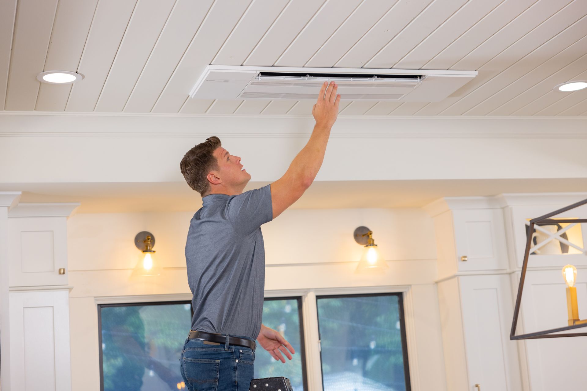 Pfluke Home Services technician on a step stool reaching to adjust ceiling air vent in a white room.