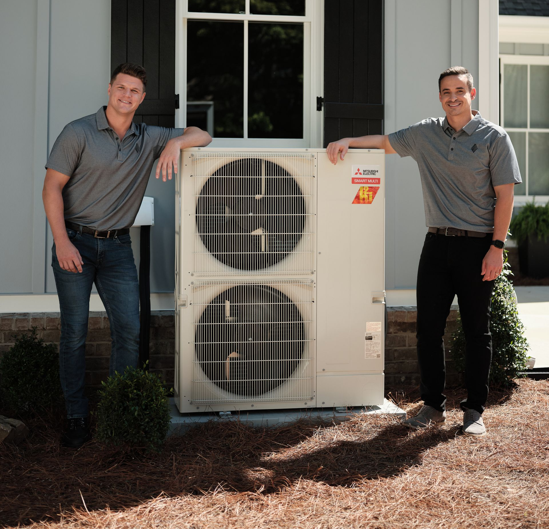Two Pfluke Home Services technician stand near an outdoor HVAC unit, both wearing gray polo shirts, posing in front of a house.