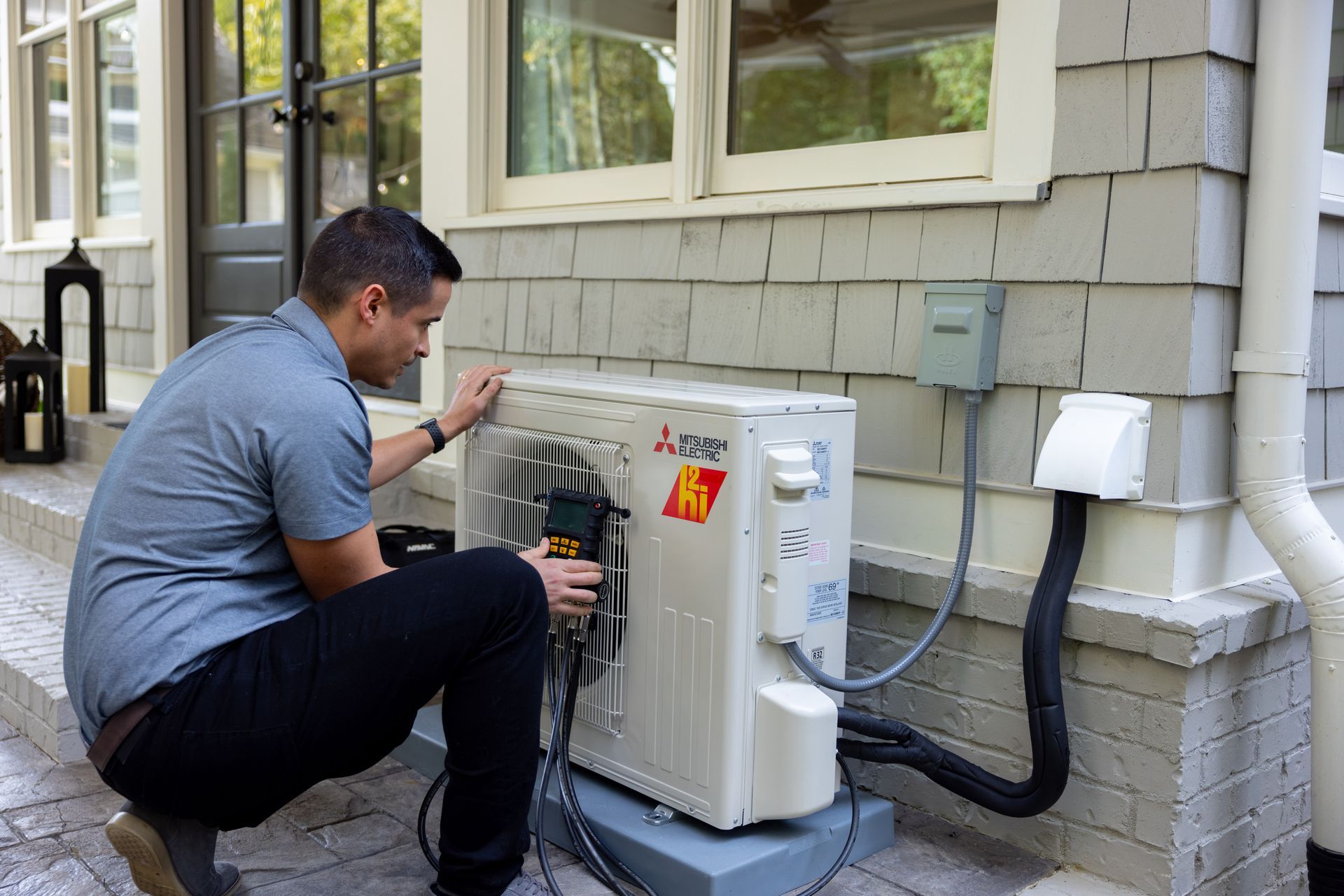 Pfluke Home Services technician installing a heat pump unit on a house exterior. Gray siding, black conduit, and electrical box visible.