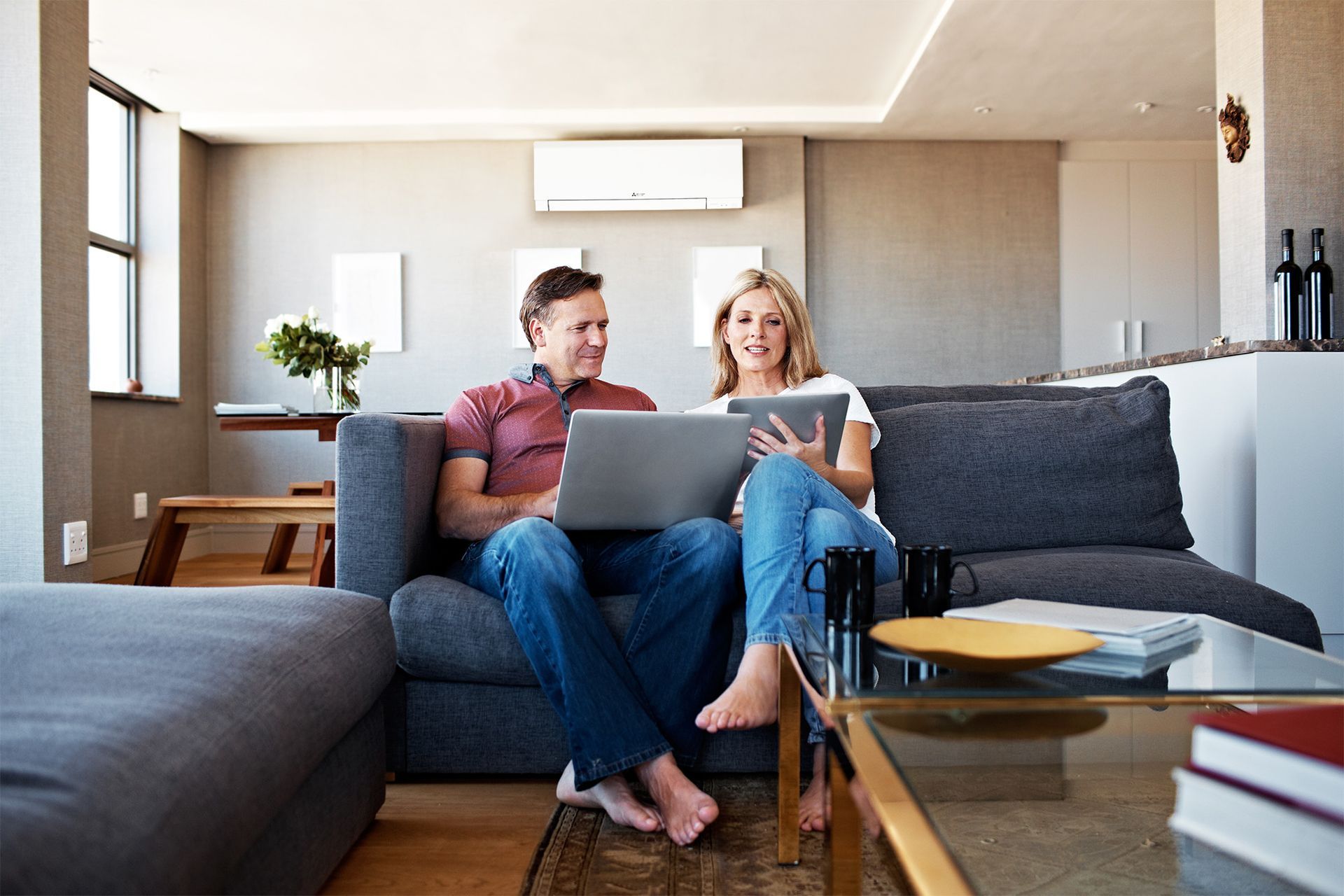 Couple on a gray sofa, man with laptop, woman with tablet, smiling in a modern living room.