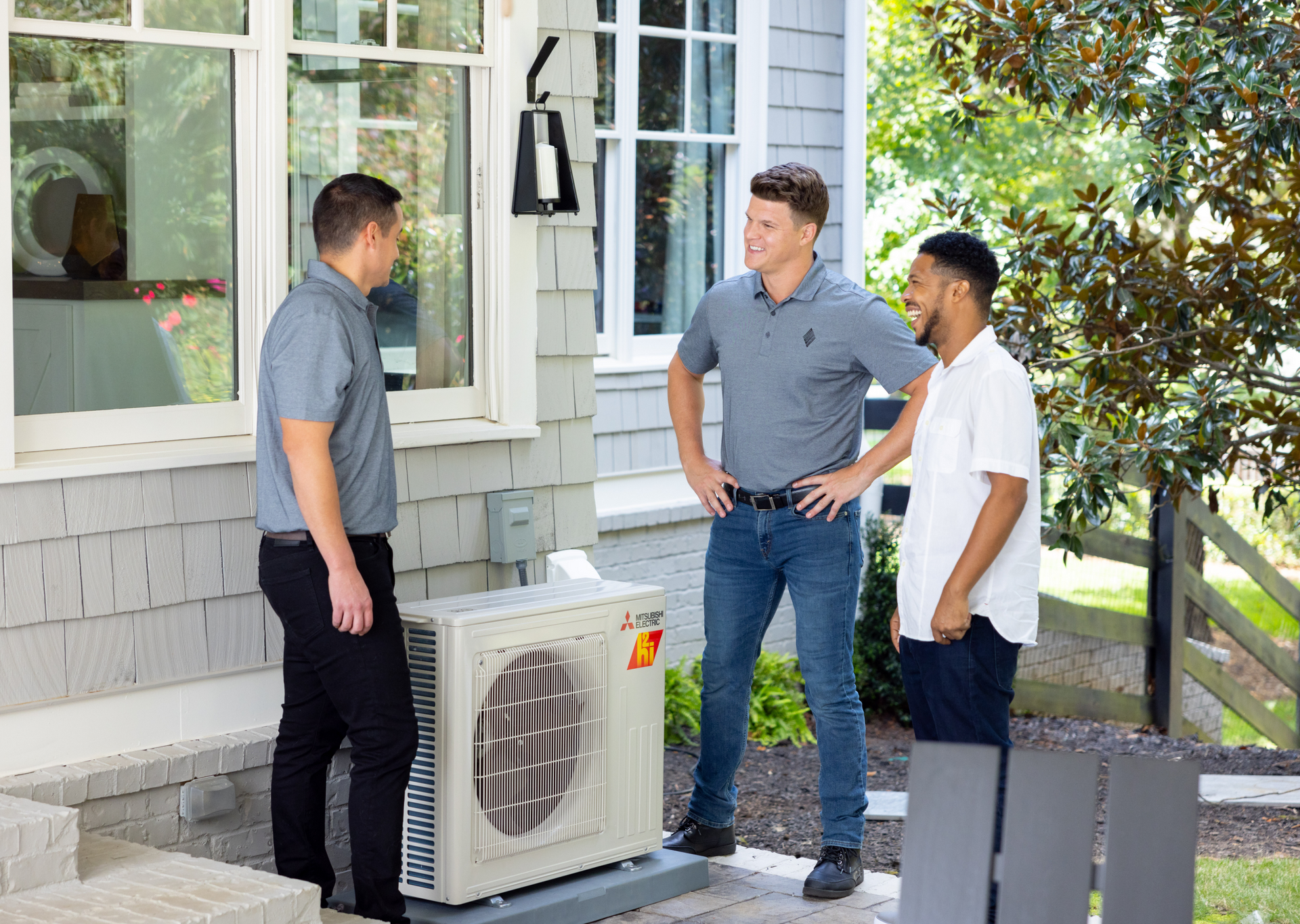 Three men by an air conditioning unit outside a house, conversing.