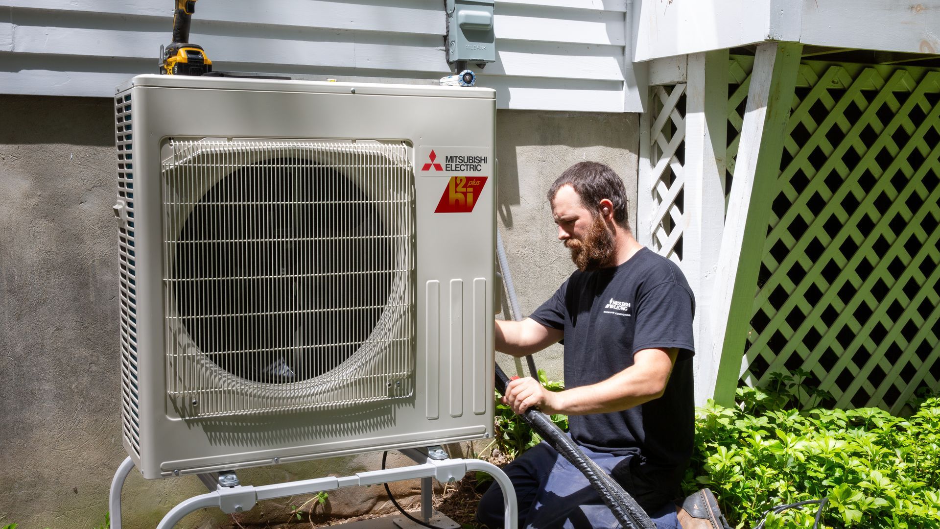 Pfluke Home Services technician servicing a Mitsubishi air conditioning unit outside a building, with a hose.