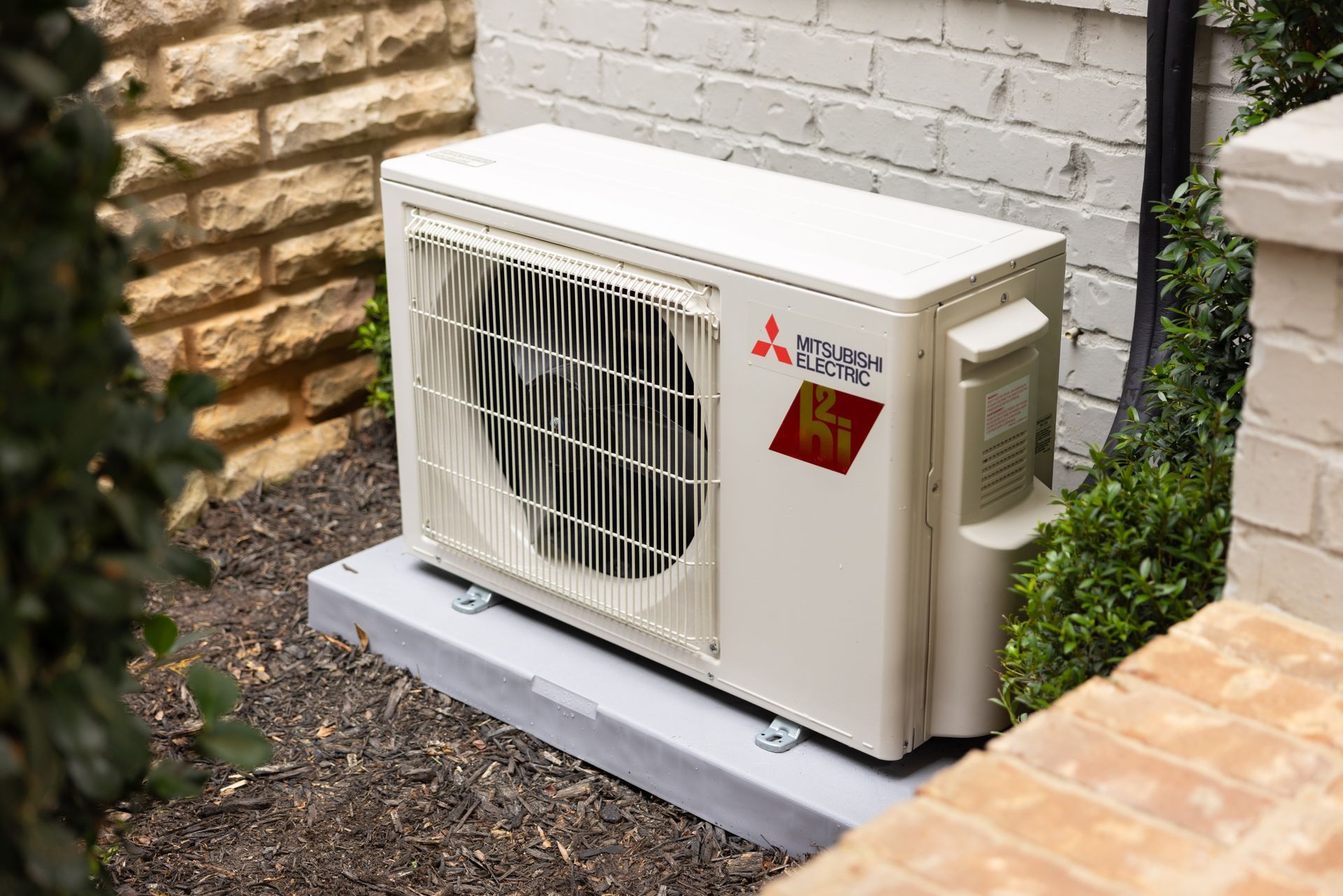 Mitsubishi heat pump unit outside near a brick wall and bushes.