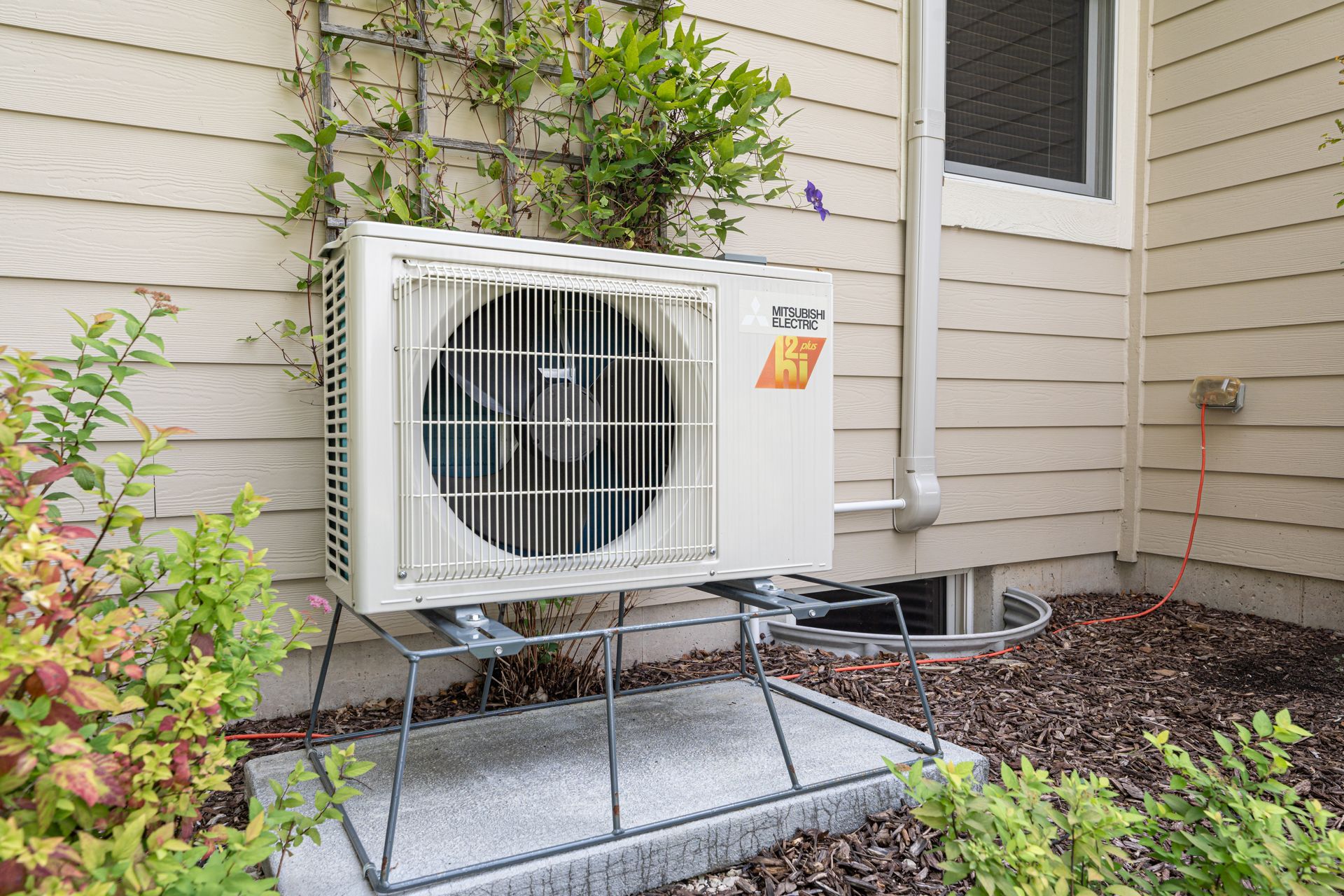 Outdoor air conditioning unit mounted on a metal stand next to a house with vegetation.