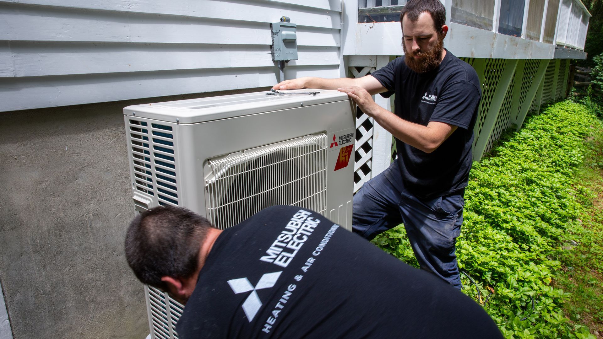 Two Pfluke Home Services technician installing a Mitsubishi Electric HVAC unit outside a house.