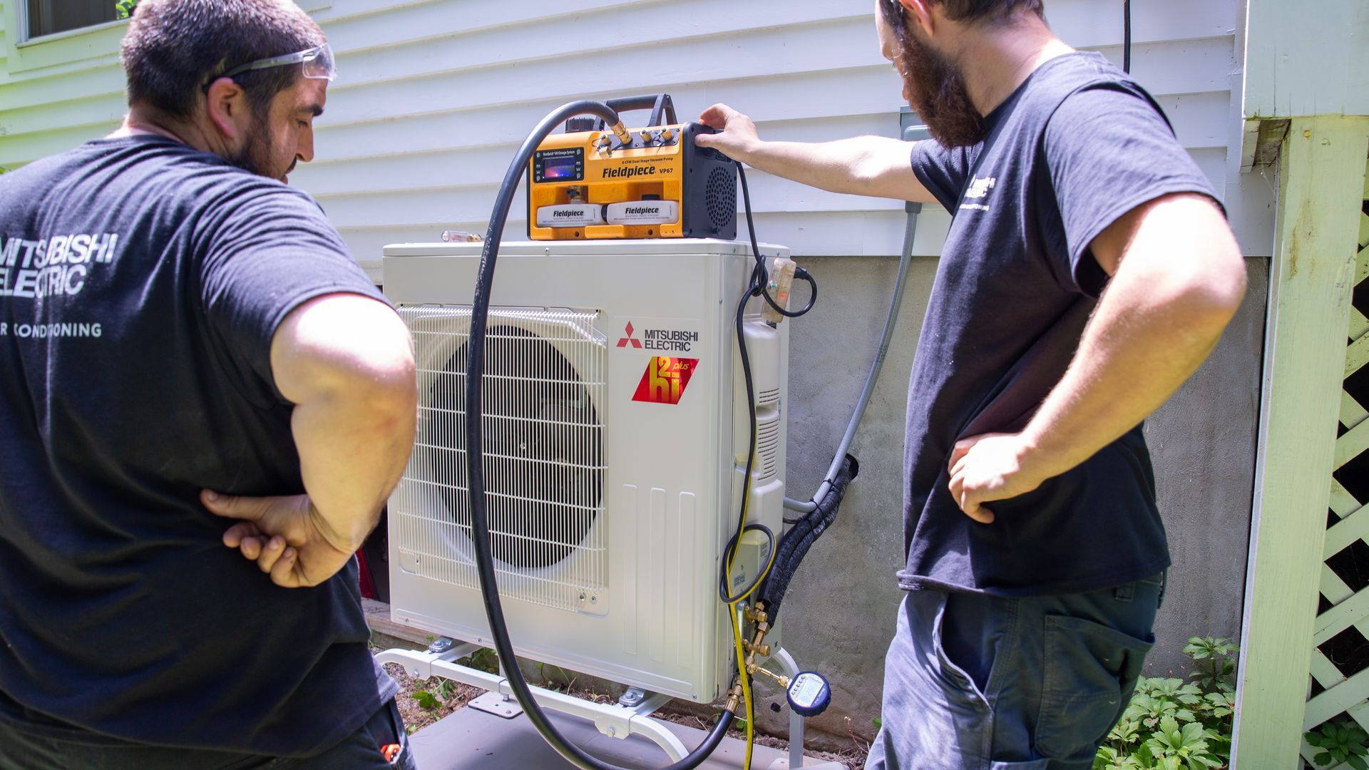 Two Pfluke Home Services technician servicing a Mitsubishi air conditioner unit outside a house.