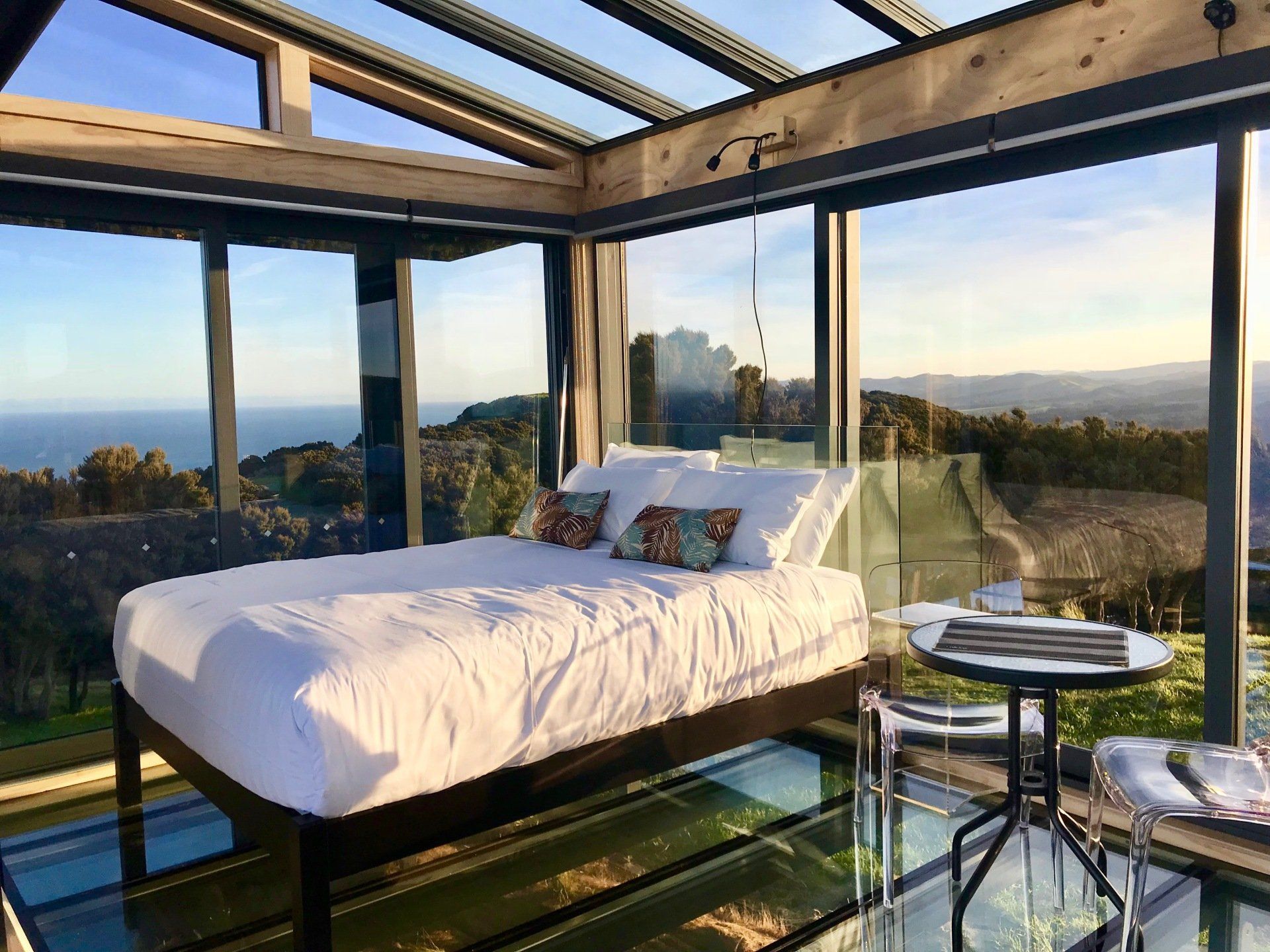 A bed is sitting in a glass room with a view of the ocean.