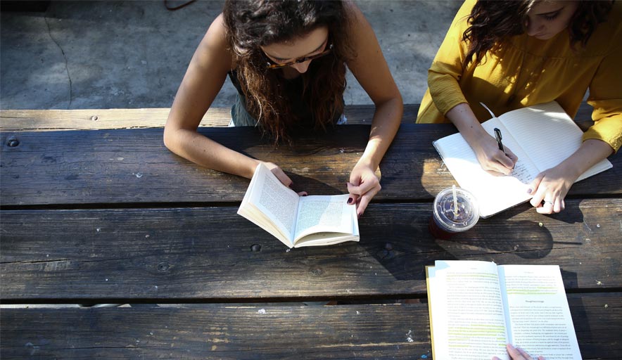 Two women are sitting at a picnic table reading books and writing in notebooks.