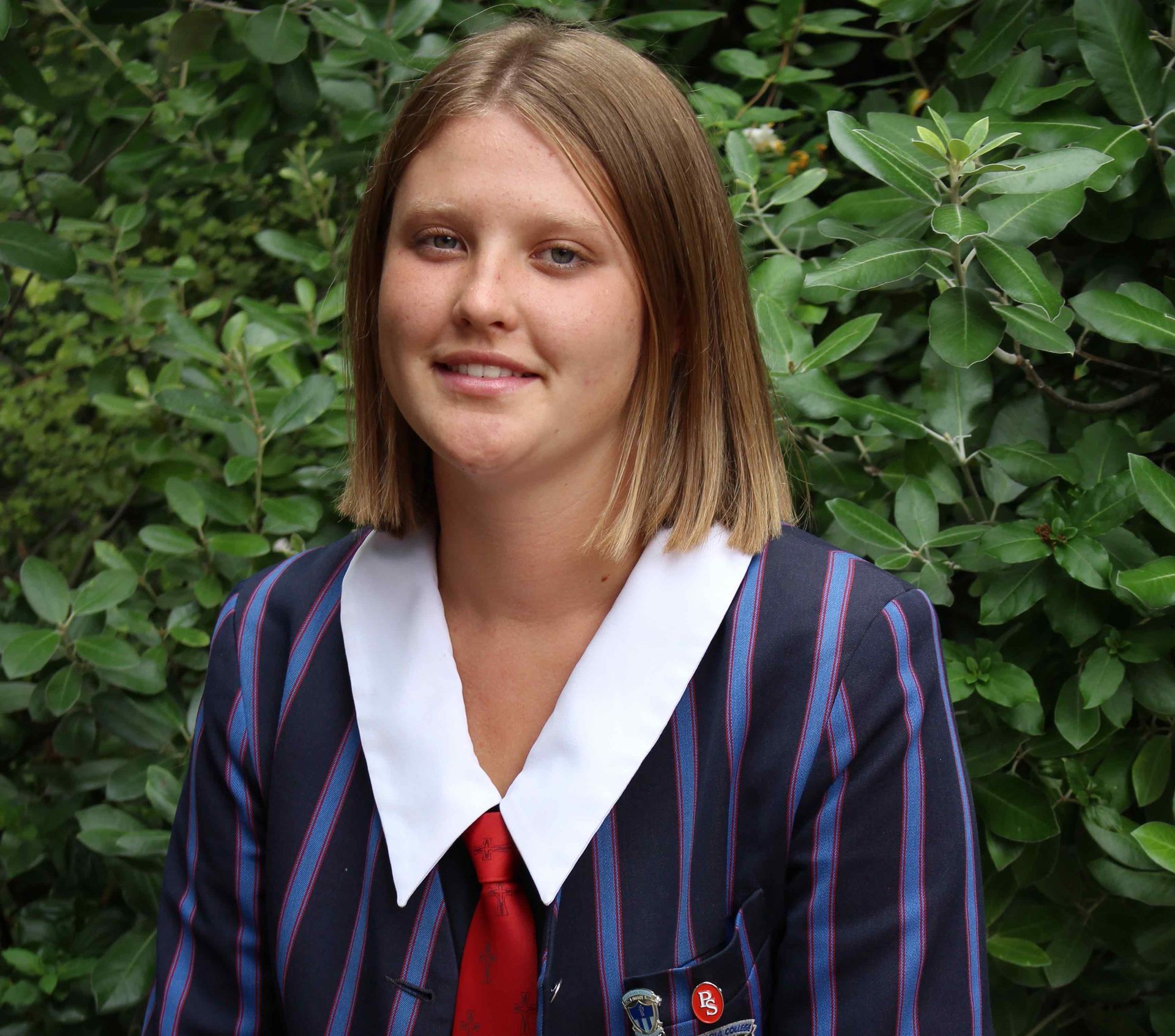 A woman wearing a striped jacket and tie is standing in front of a bush.