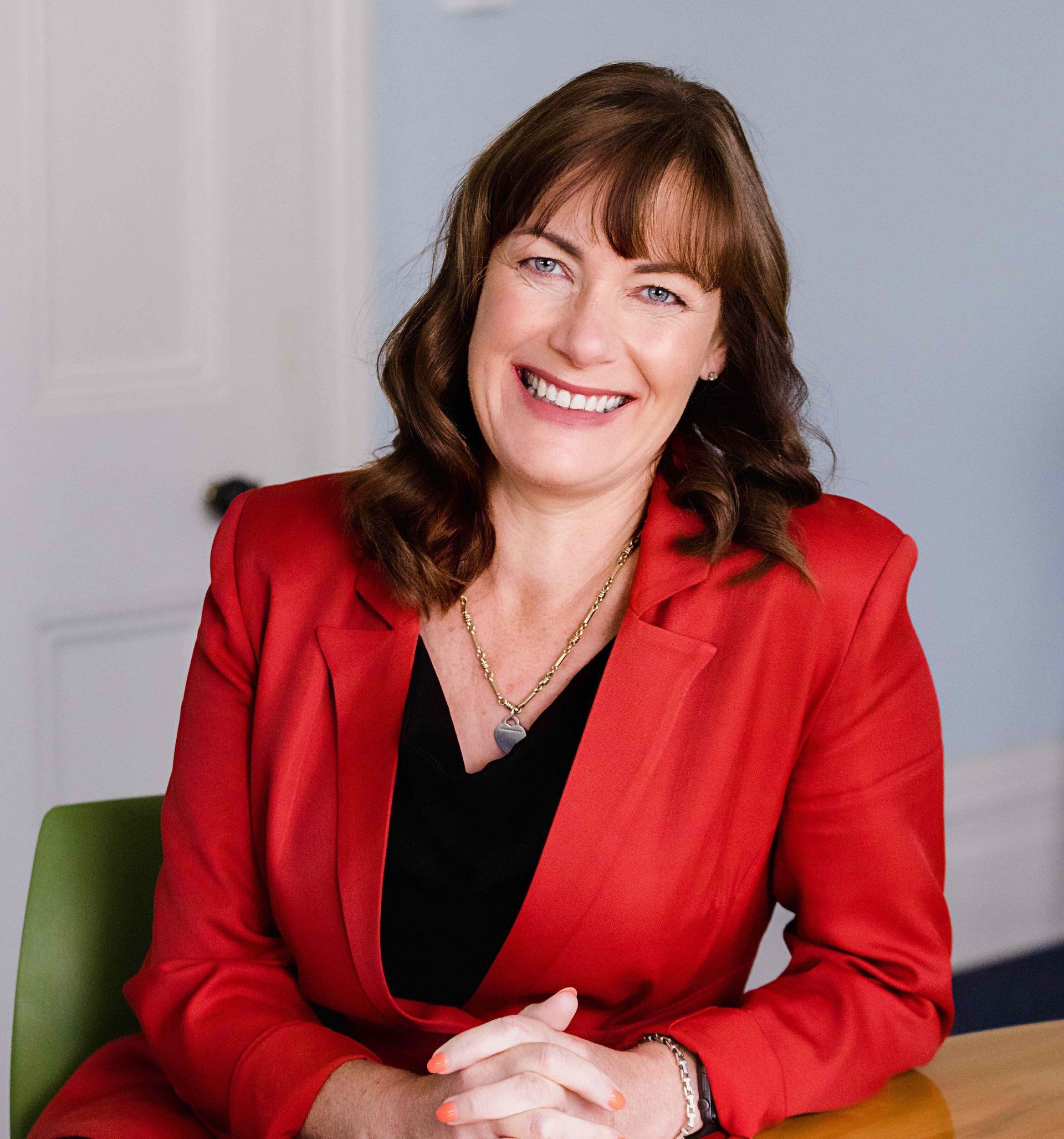 A woman in a red jacket is sitting at a table with her hands folded and smiling.