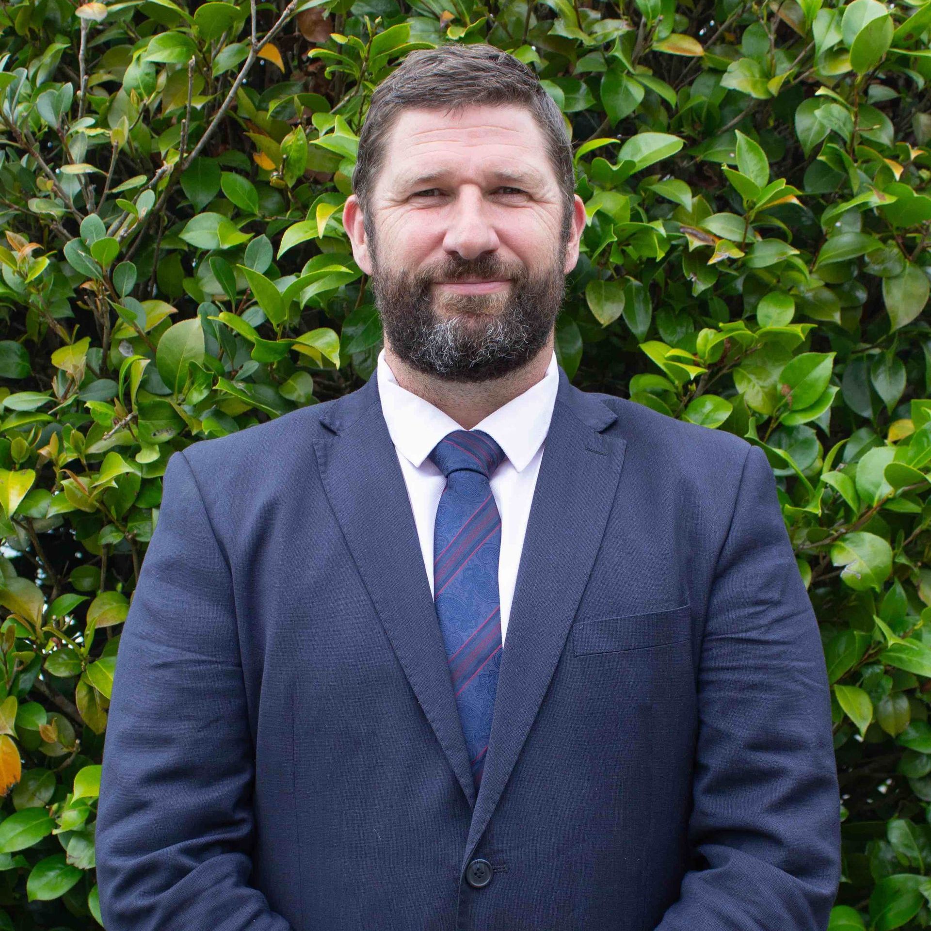 A man in a suit and tie is standing in front of a bush.