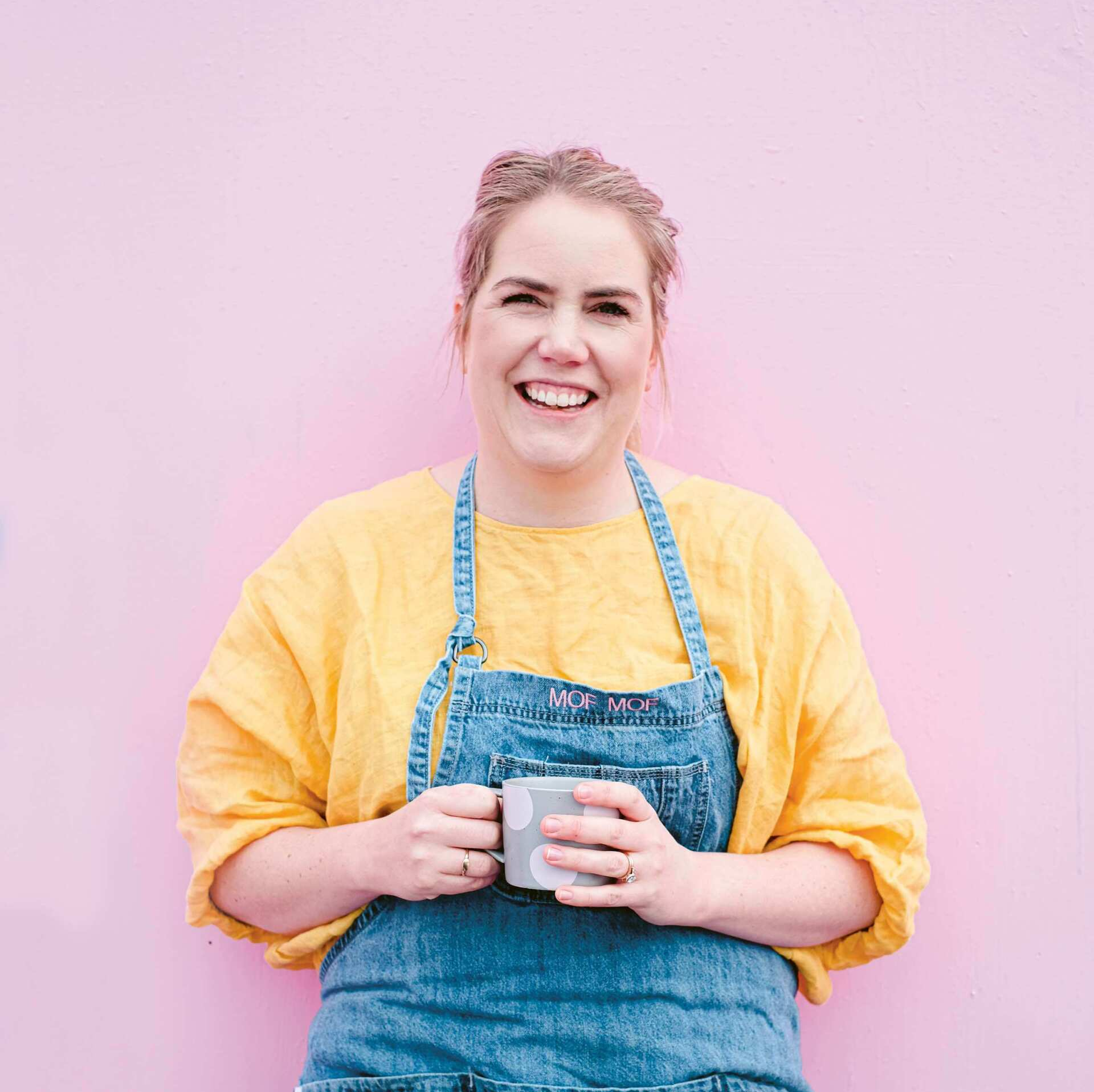 A woman in an apron is holding a cup of coffee and smiling.