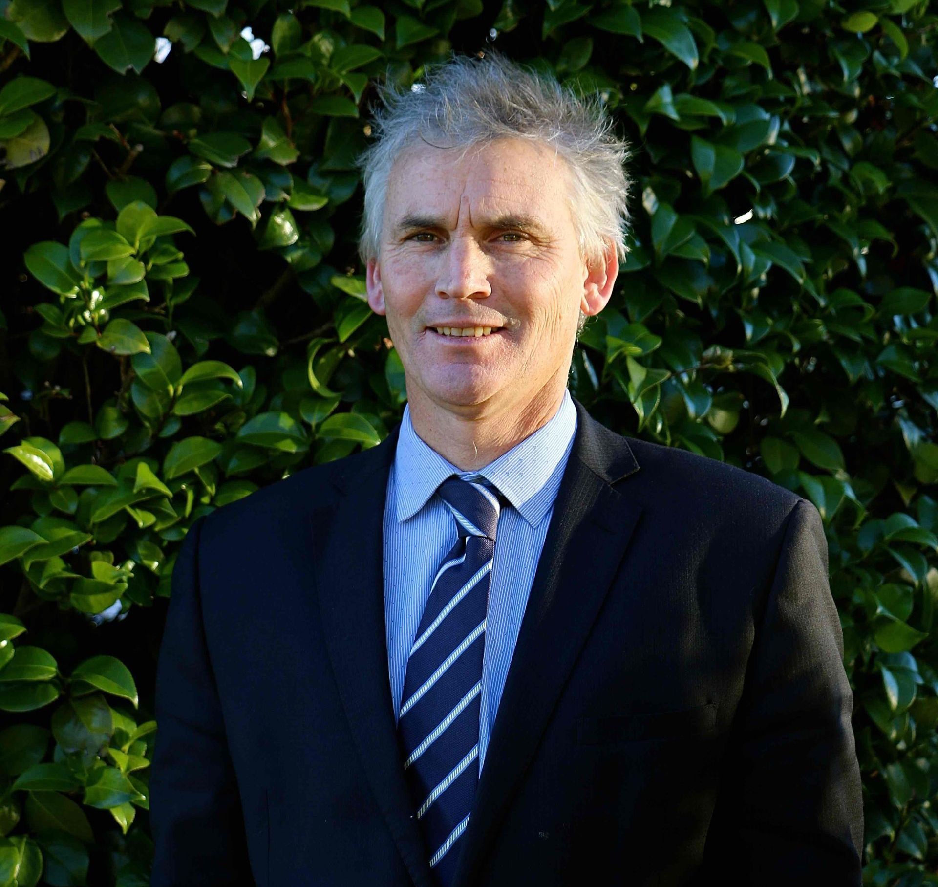 A man in a suit and tie is standing in front of a bush.